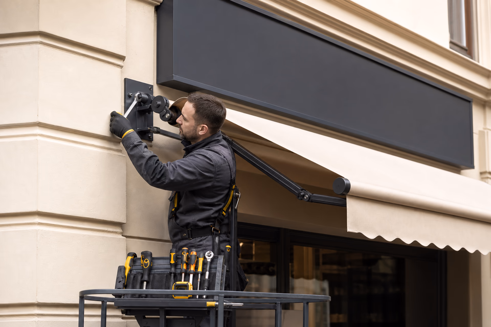 Awning installer tightening a bracket on a cream storefront canopy beneath a blank charcoal sign panel